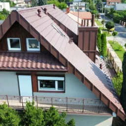 Dom jednorodzinny z brązowym dachem z blachodachówki, widok z góry na dach, komin i okna dachowe, otoczony zielenią i innymi budynkami.