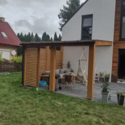 Nowoczesna pergola z drewnianymi lamelami i czarną ramą, przylegająca do białej ściany domu z drewnianą okładziną, z wiszącym hamakiem i meblami ogrodowymi na szarym tarasie