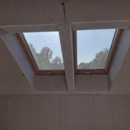 Dwa okna dachowe zamontowane w poddaszu wykończonym płytami kartonowo-gipsowymi, widoczne kable wystające z sufitu, widok na drzewa za oknem.