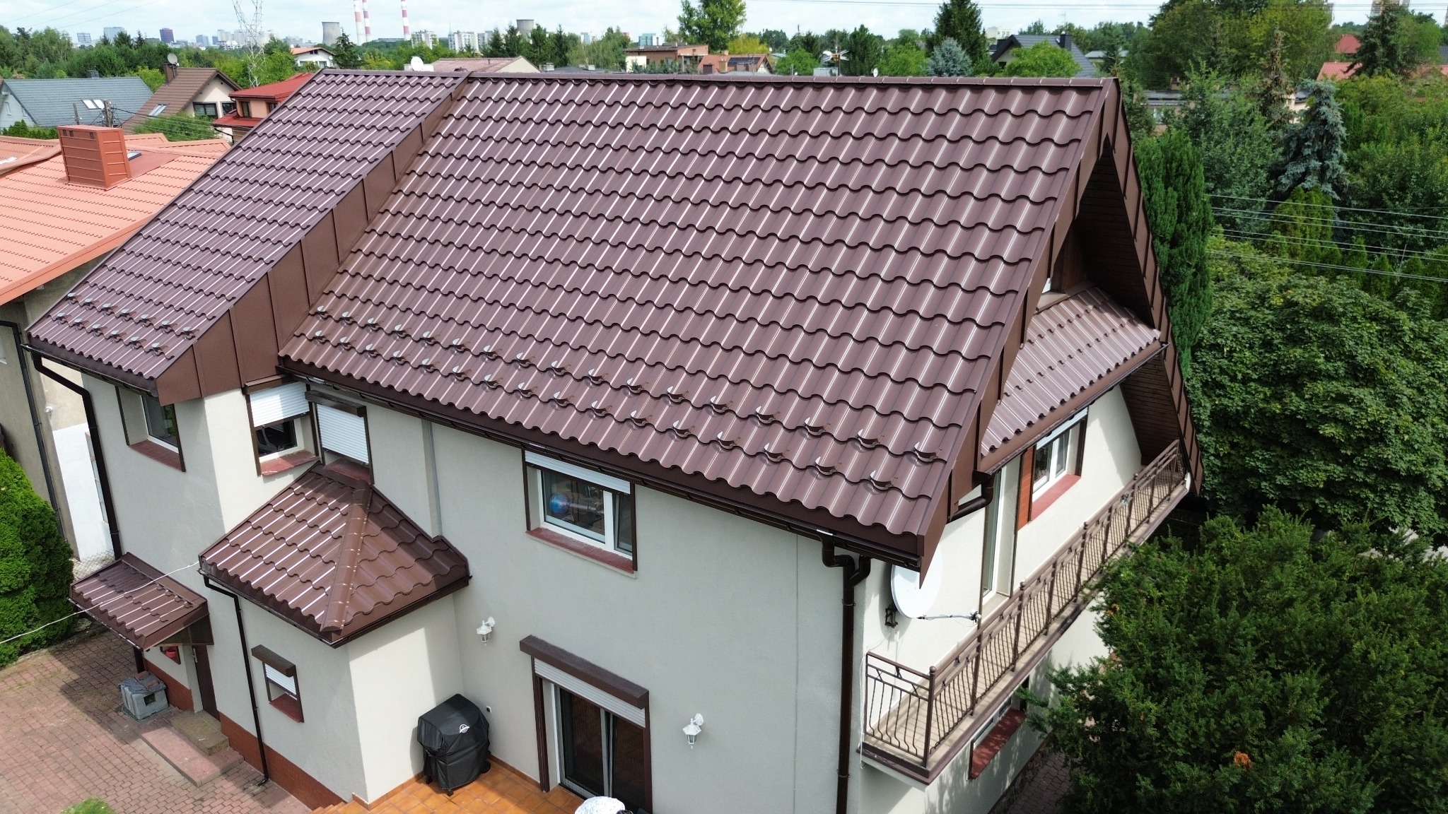 Dom jednorodzinny z brązowym dachem z blachodachówki modułowej, widok z góry, z widocznymi detalami takimi jak rynny, okna z roletami zewnętrznymi i balkon z ażurową balustradą.