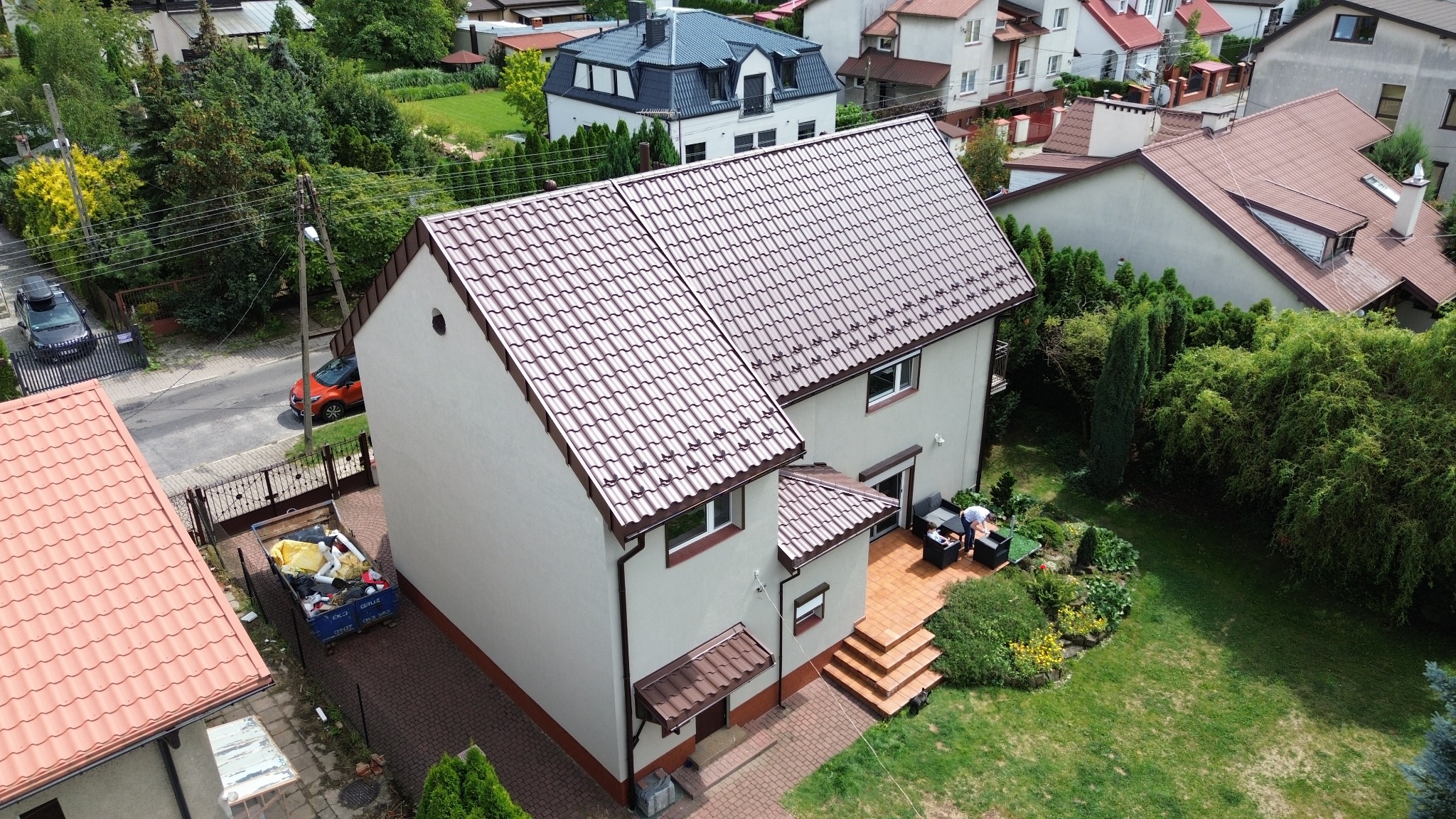 Dwupiętrowy dom z beżową elewacją i brązowym dachem z dachówki, widok z góry, z ogrodem i patio na tyłach, na którym stoi zestaw mebli ogrodowych.