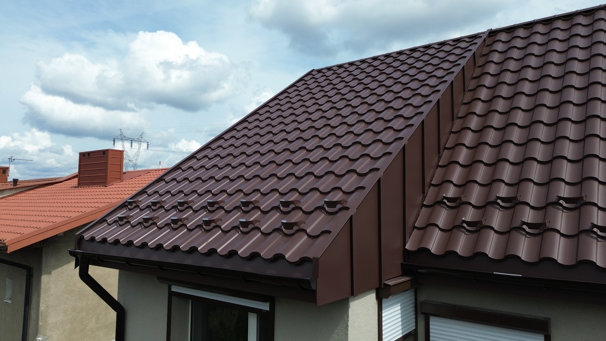 Ciemnobrązowe, metalowe pokrycie dachowe z widocznymi odpowietrznikami, zamontowane na budynku z jasną elewacją i oknami z roletami zewnętrznymi, na tle błękitnego nieba z chmurami.