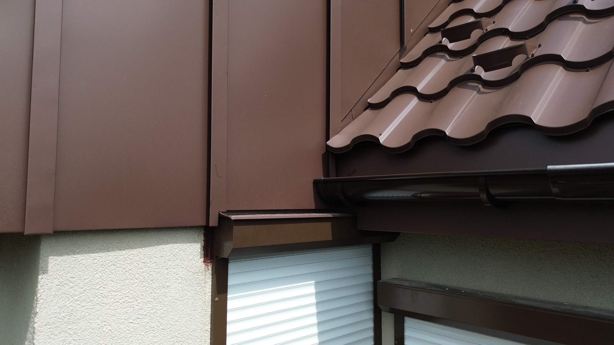 Fragment budynku z brązową elewacją panelową i dachówką ceramiczną w odcieniach brązu, widoczne rynny i rolety zewnętrzne.