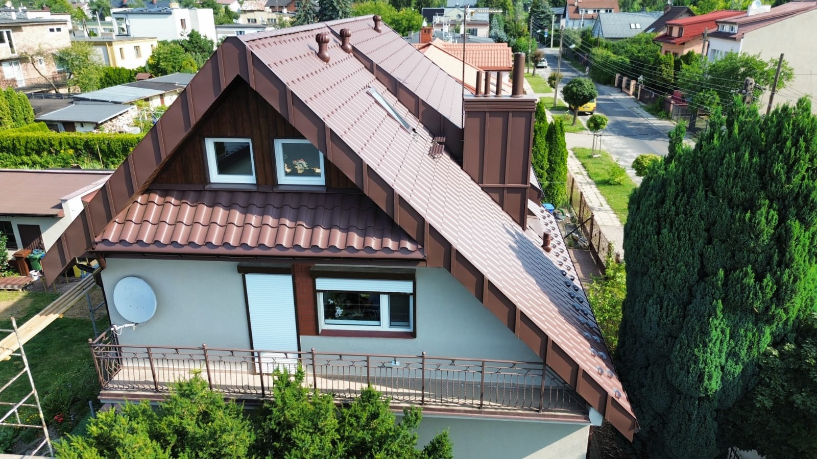 Dom jednorodzinny z brązowym dachem z blachodachówki, widok z góry na dach, komin i okna dachowe, otoczony zielenią i innymi budynkami.