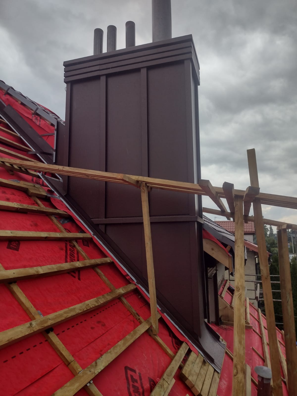 Montaż brązowego komina wentylacyjnego z trzema wylotami na dachu z czerwoną membraną i drewnianą konstrukcją, widok z dołu pod kątem w pochmurny dzień.