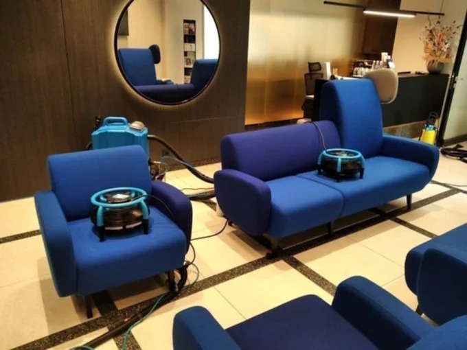 Pranie niebieskiej sofy i foteli w biurze. Na meblach stoją urządzenia do ekstrakcyjnego czyszczenia tapicerki. W tle widoczne lustro i recepcja.