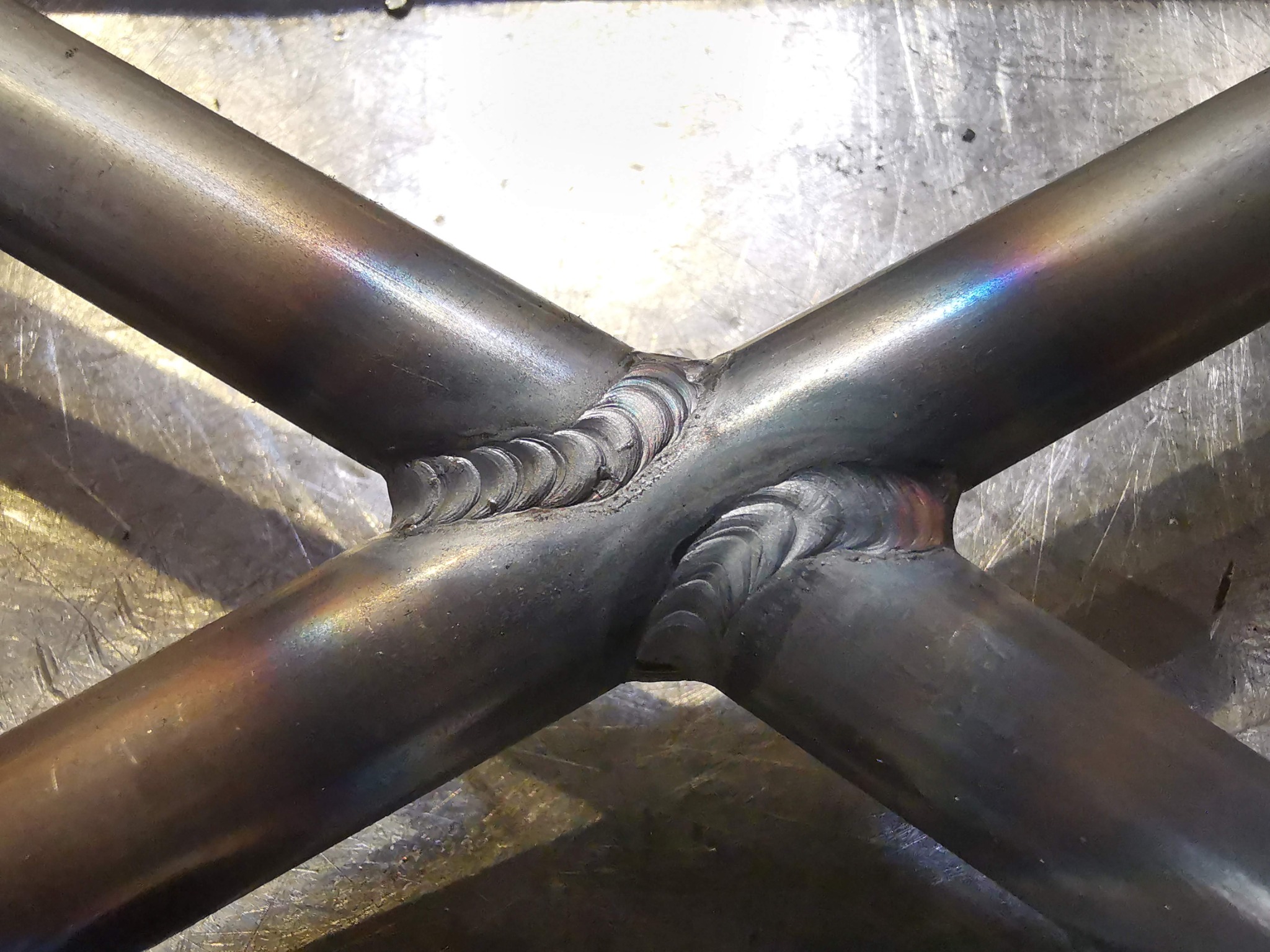 Spoina krzyżowa dwóch metalowych rur, widoczne ślady spawania TIG i opalizujące zabarwienie metalu w miejscu spawu na tle metalowej powierzchni roboczej.