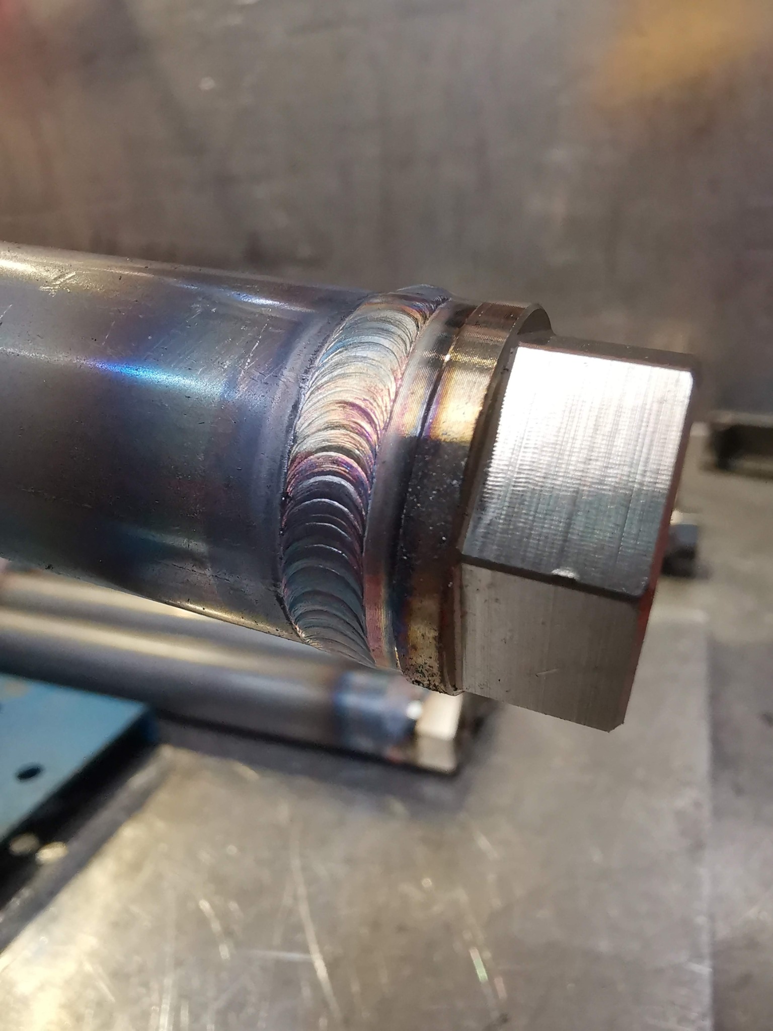 Szczegółowe zbliżenie spoiny łączącej metalową rurę z sześciokątną nakrętką, widoczne tęczowe przebarwienia na spoinie, ślady obróbki na nakrętce.