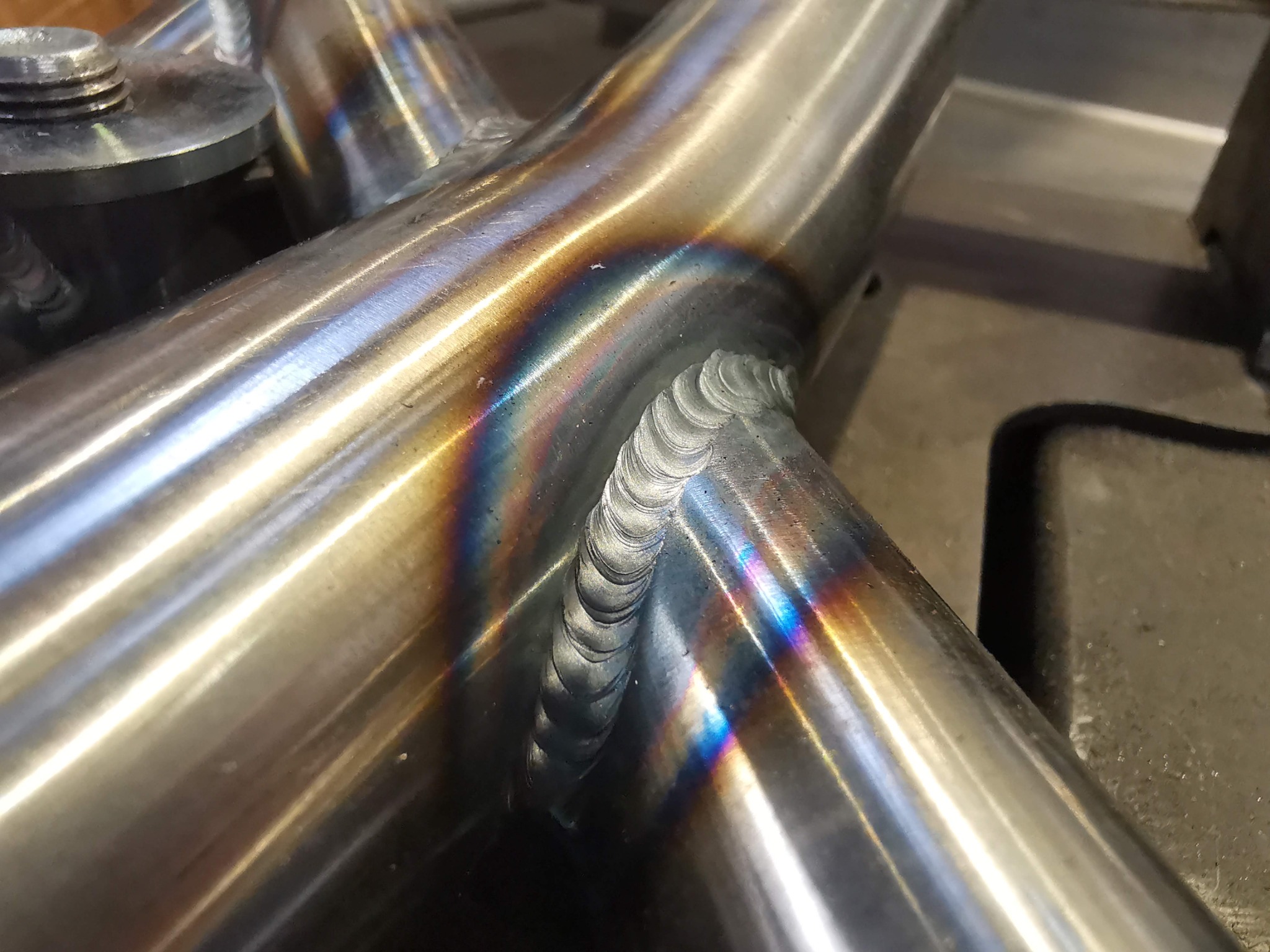 Szczegółowe zbliżenie na spoinę TIG łączącą trzy rury ze stali nierdzewnej, widoczne tęczowe przebarwienia termiczne.