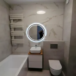 Nowoczesna łazienka z białymi płytkami imitującymi marmur, okrągłym podświetlanym lustrem, białą umywalką nablatową na szafce z drewnianym blatem, wiszącą toaletą i grzejnikiem drabinkowym.