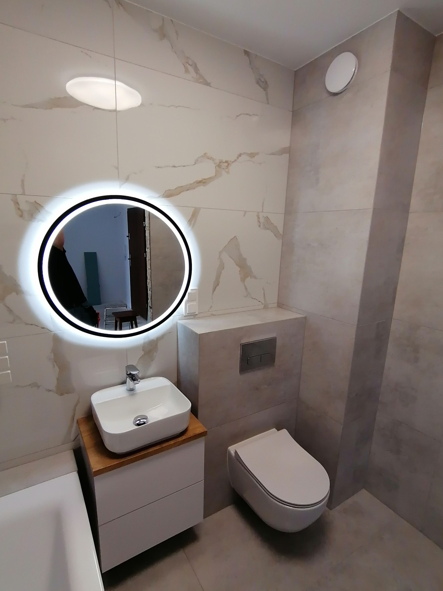 Nowoczesna łazienka z marmurowymi płytkami, okrągłym lustrem z podświetleniem LED, wiszącą toaletą i umywalką nablatową na szafce z drewnianym blatem.