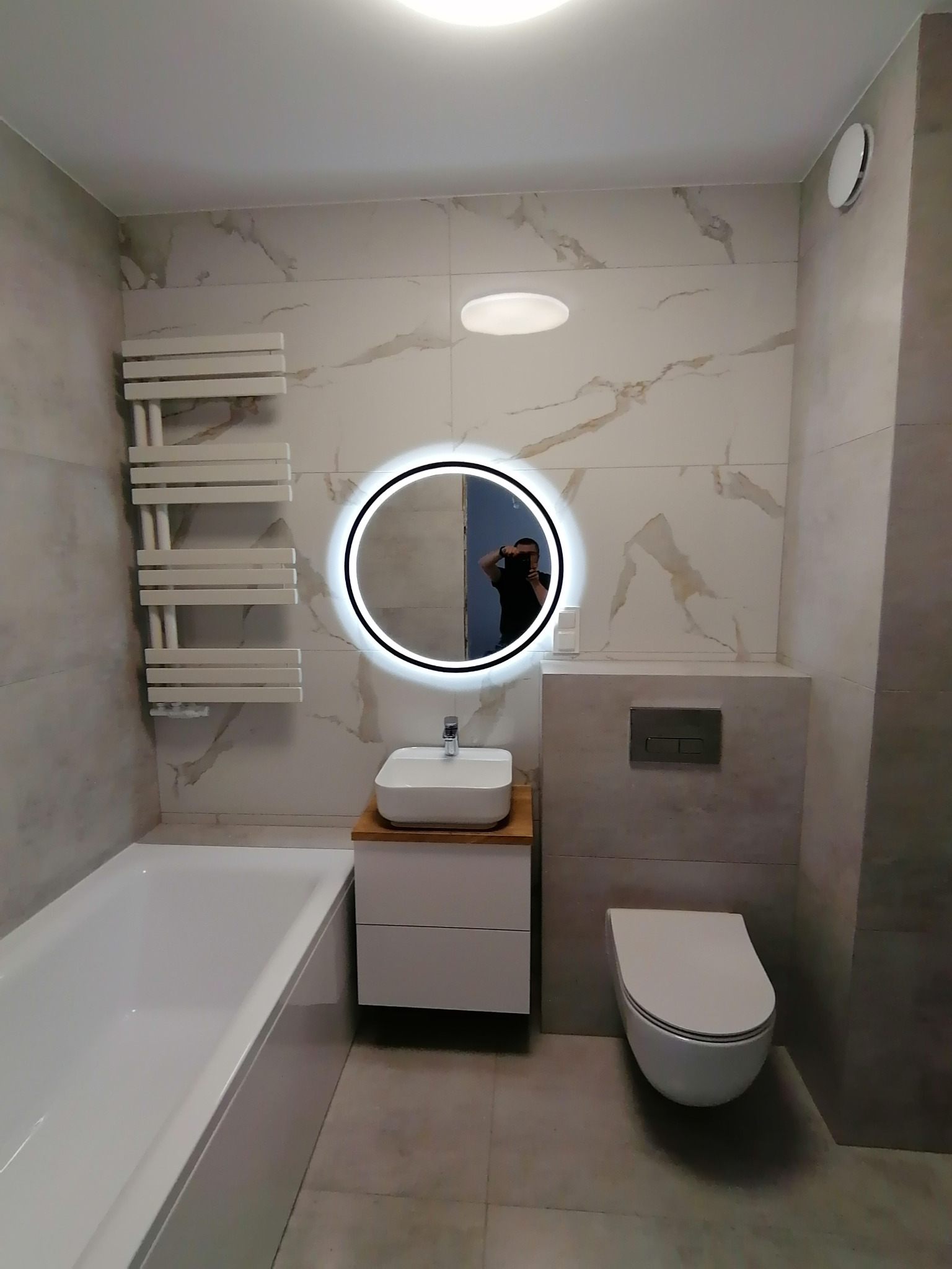 Nowoczesna łazienka z białymi płytkami imitującymi marmur, okrągłym podświetlanym lustrem, białą umywalką nablatową na szafce z drewnianym blatem, wiszącą toaletą i grzejnikiem drabinkowym.