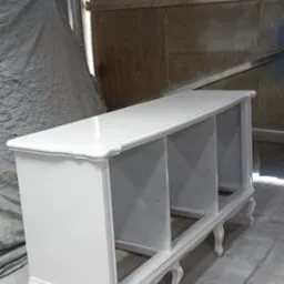 Biała, odnowiona komoda z trzema otwartymi przegródkami i dekoracyjnymi nóżkami w stylu retro, stojąca w warsztacie, widoczne ślady farby na podłodze.