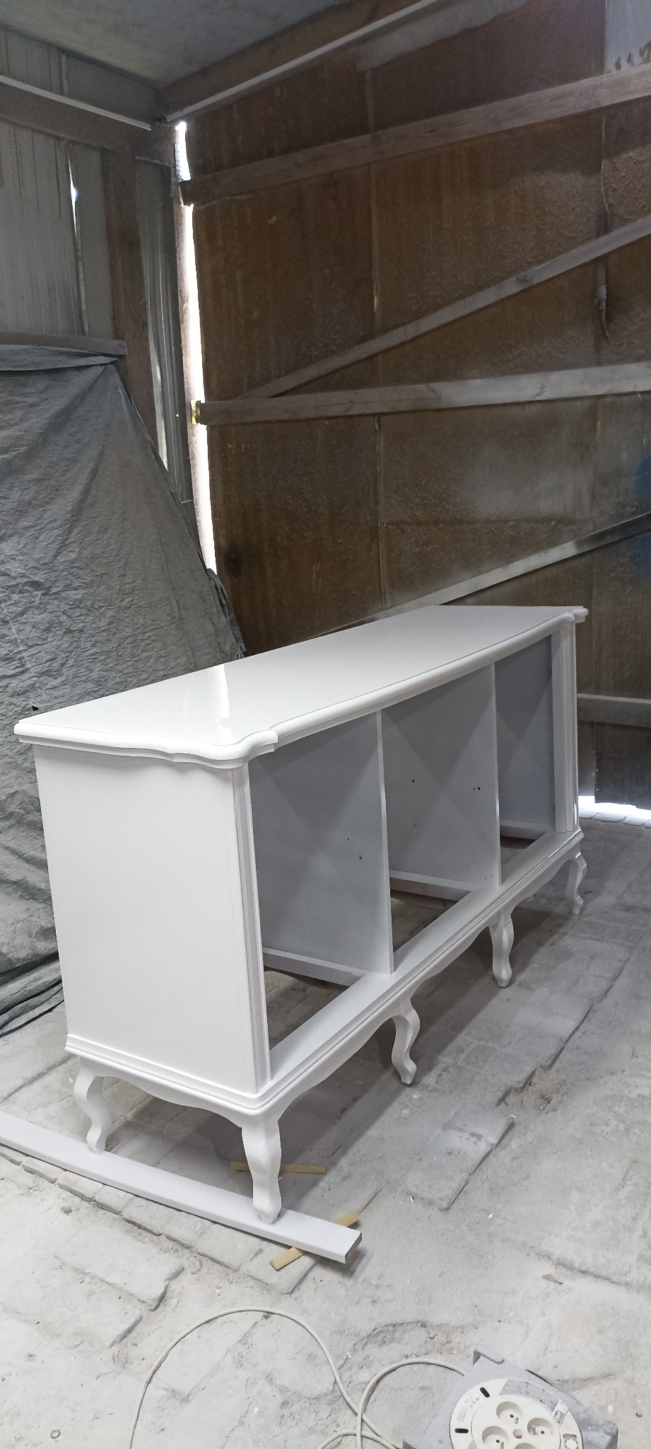 Biała, odnowiona komoda z trzema otwartymi przegródkami i dekoracyjnymi nóżkami w stylu retro, stojąca w warsztacie, widoczne ślady farby na podłodze.