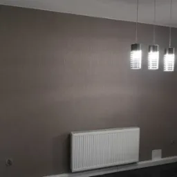 Pokój z szarobrązową ścianą o delikatnej fakturze, białym grzejnikiem panelowym, trzema nowoczesnymi lampami wiszącymi i czarną podłogą. Na ścianie wisi obraz z mapą świata w białej ramie.