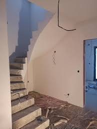 Wnętrze w trakcie prac wykończeniowych: surowe schody betonowe prowadzą na górę, ściany pokryte świeżą gładzią, widoczne wystające kable elektryczne i przygotowane podłoże pod posadzkę, otwarte...