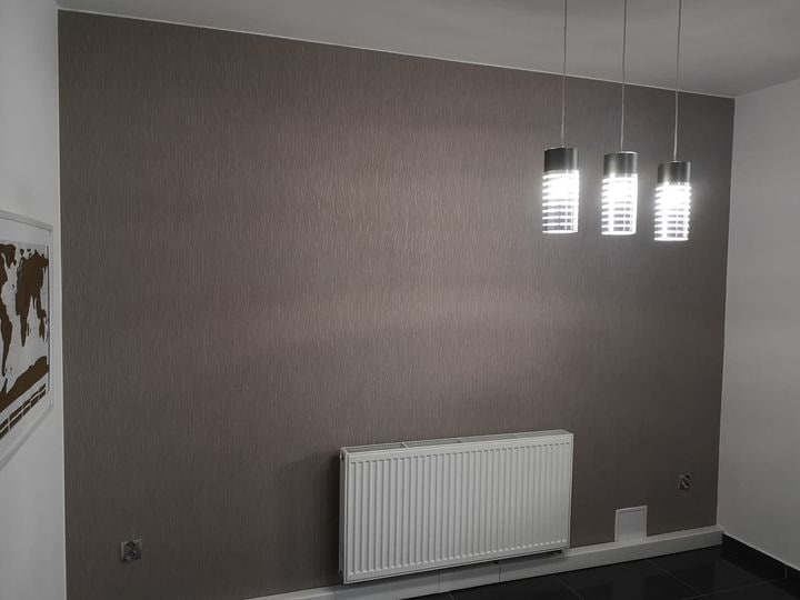 Pokój z szarobrązową ścianą o delikatnej fakturze, białym grzejnikiem panelowym, trzema nowoczesnymi lampami wiszącymi i czarną podłogą. Na ścianie wisi obraz z mapą świata w białej ramie.