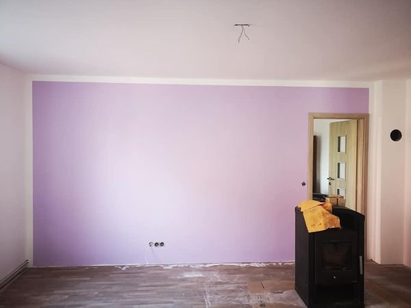 Ściana pomalowana na fioletowo z widocznymi gniazdkami elektrycznymi, fragmentem podłogi zabezpieczonej kartonem i piecem typu koza przy ścianie, w tle otwarte drzwi.