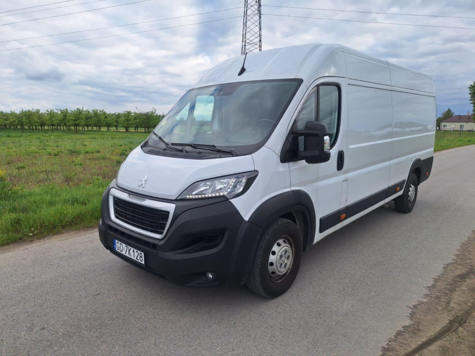 Biały bus Peugeot Boxer z numerem rejestracyjnym GD 7K128 stoi na asfalcie, w tle pole z drzewami i słup wysokiego napięcia pod pochmurnym niebem.