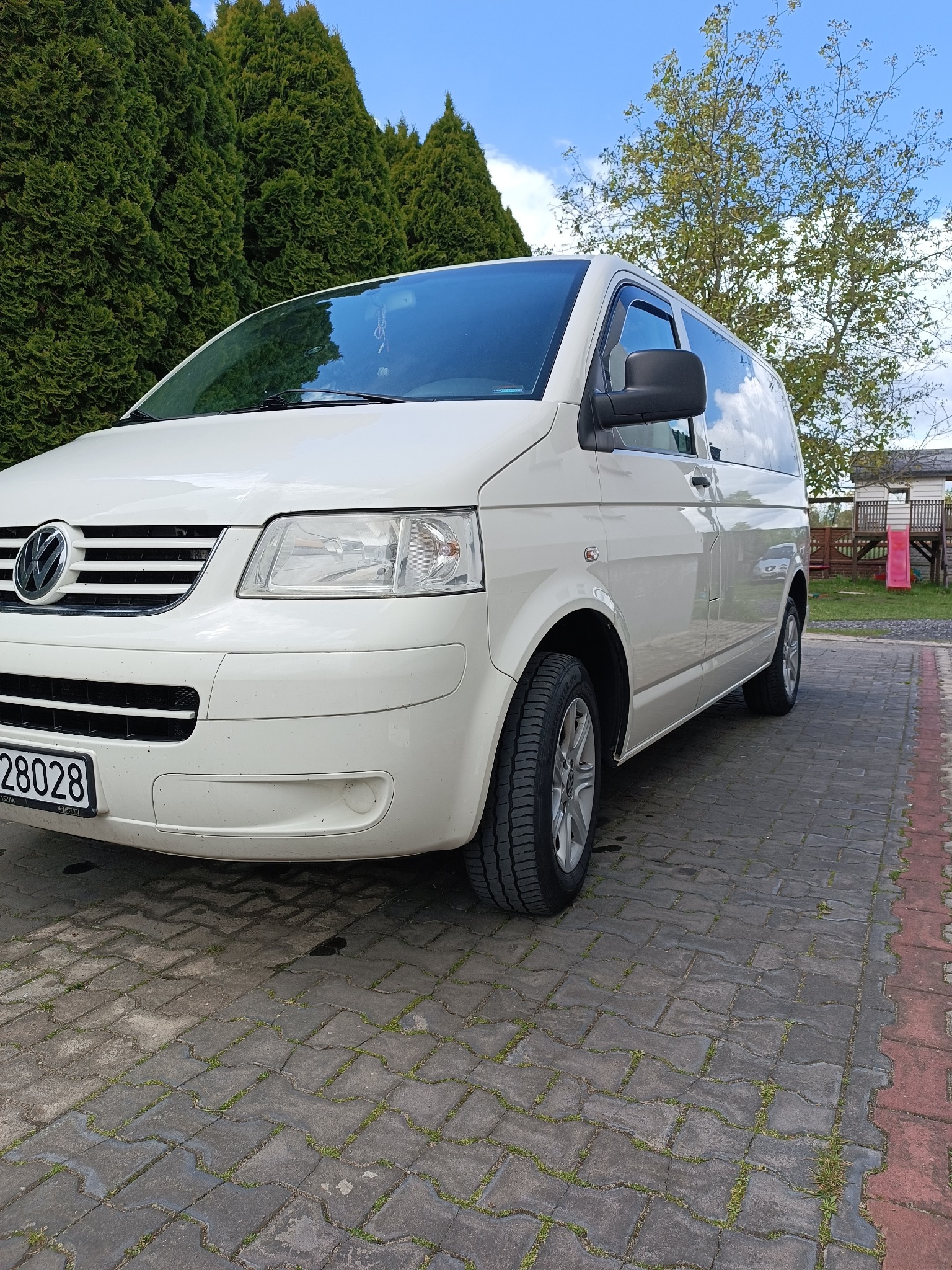 Biały Volkswagen Transporter T5 z widocznym numerem rejestracyjnym, zaparkowany na kostce brukowej, z zielonymi drzewami w tle i fragmentem placu zabaw.