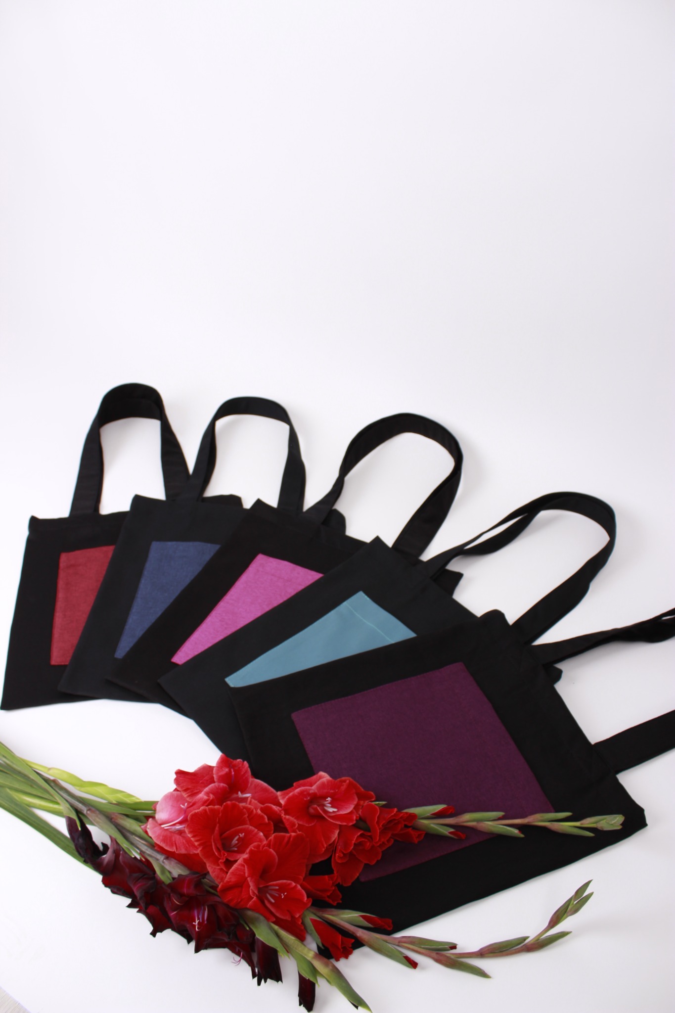 Pięć czarnych toreb materiałowych z kolorowymi kwadratami, ułożonych obok siebie, ozdobionych czerwonymi mieczykami na białym tle.