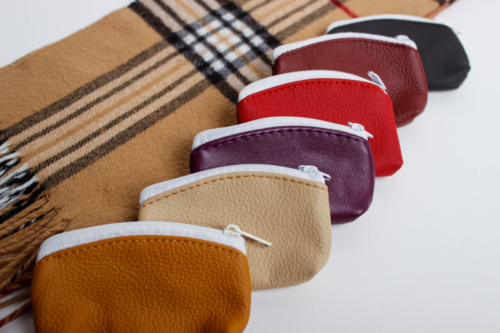 Rząd małych, kolorowych portmonetek ze skóry ekologicznej z zamkami błyskawicznymi, ułożonych na tle kaszmirowego szala w kratę.