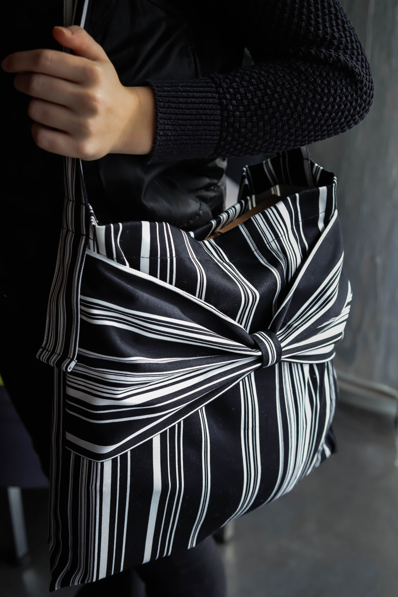 Osoba trzyma czarno-białą torbę na ramię z dekoracyjnym węzłem, wykonaną z tkaniny w pionowe paski.