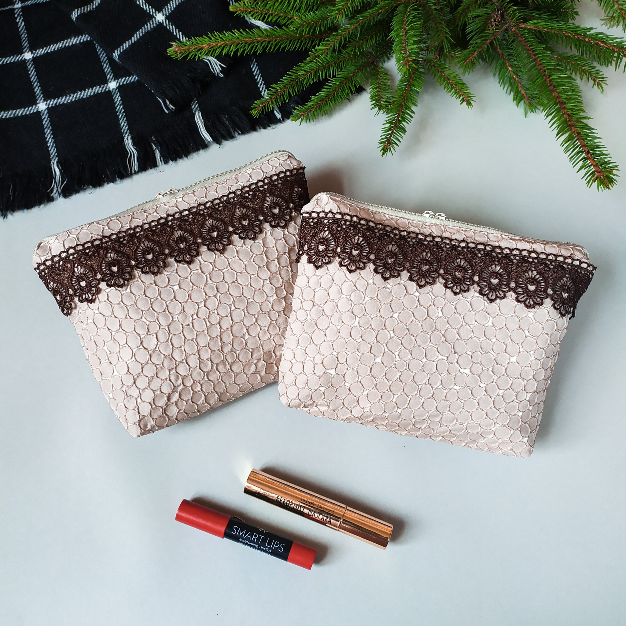 Dwie beżowe kosmetyczki z brązową koronką, leżące obok kosmetyków i gałązki świerku na białym tle.