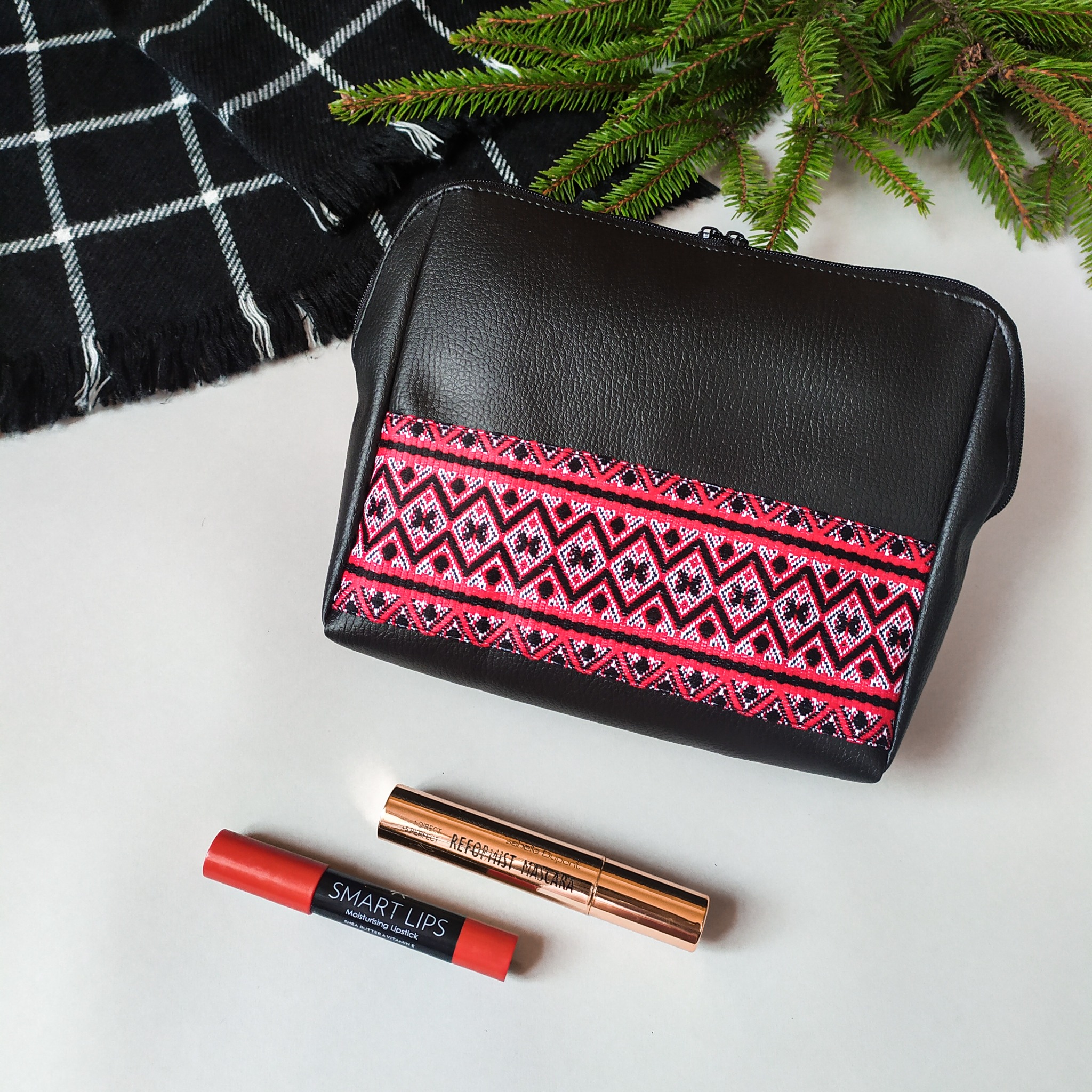 Czarna, skórzana kosmetyczka z etnicznym wzorem, leżąca na białym tle obok czarno-białego szala w kratę, gałązki świerku i kosmetyków do makijażu.
