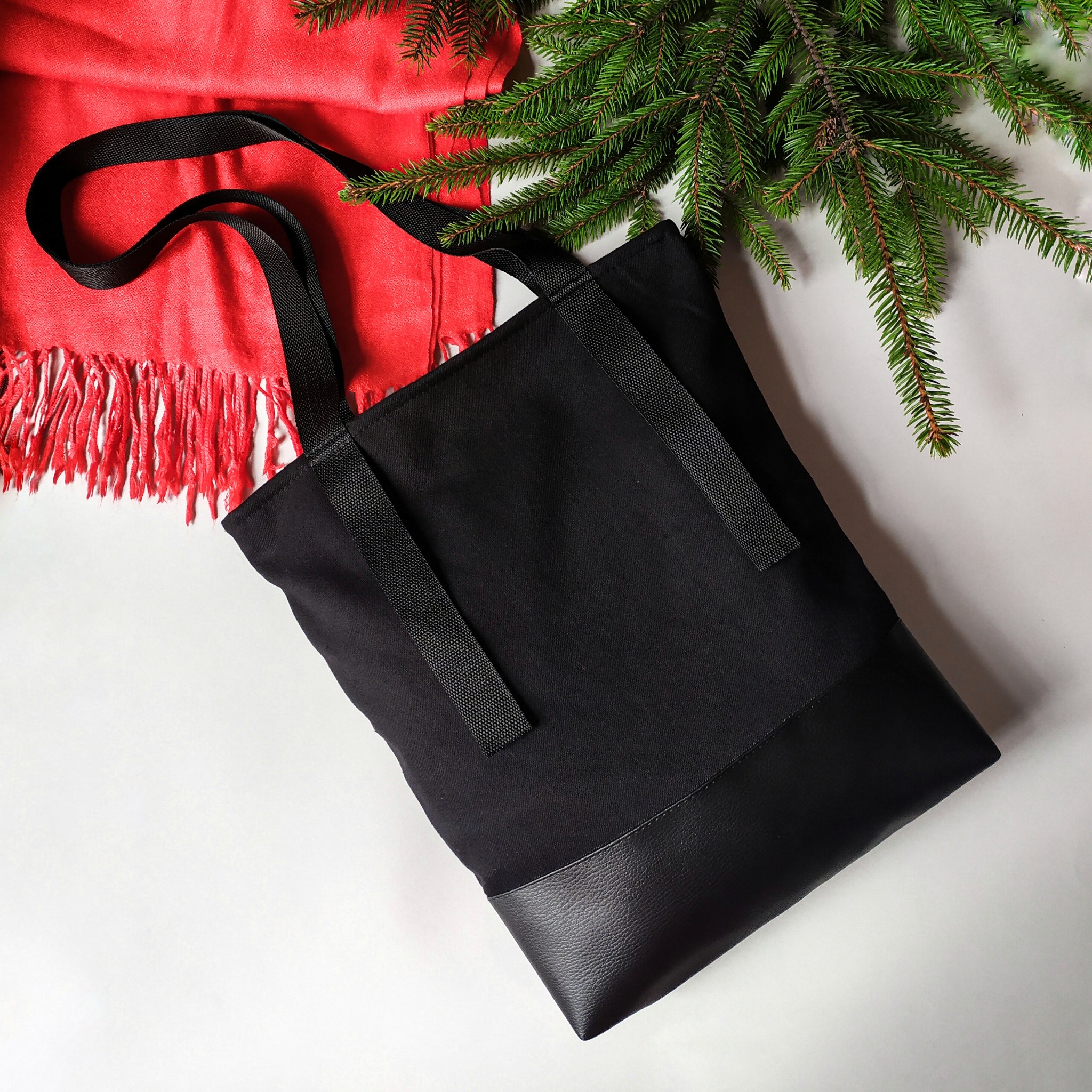 Czarna torba shopper z tkaniny i ekoskóry, na białym tle z czerwoną chustą z frędzlami i gałązką świerku, prezentowana z bliska.