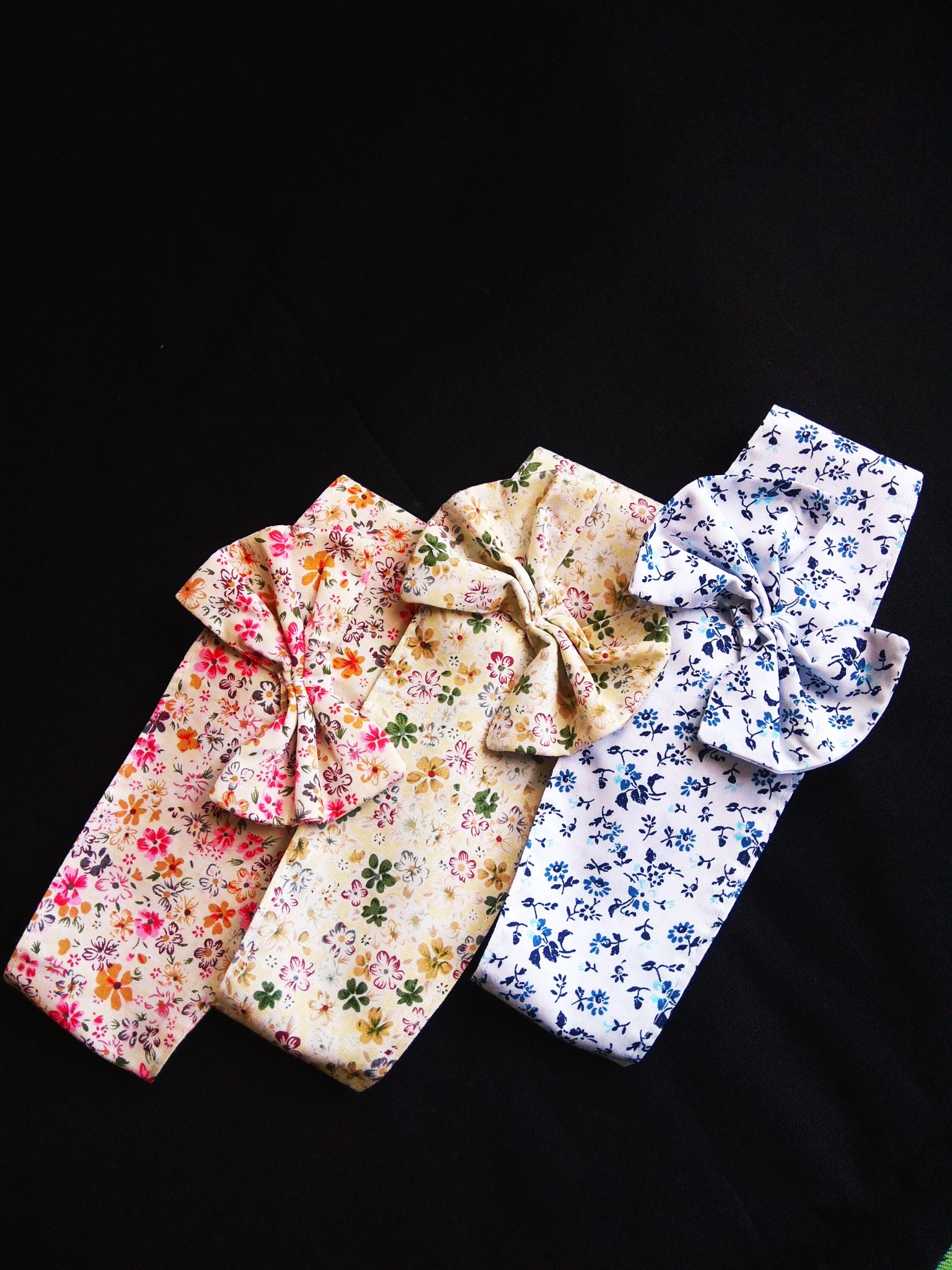 Trzy opaski do włosów z kokardami, każda w innym kwiecistym wzorze, ułożone ukośnie na czarnym tle.