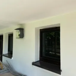 Świeżo pomalowana elewacja z oknami w ciemnych ramach i nowoczesną lampą zewnętrzną, widoczne złożone krzesła ogrodowe w tle.