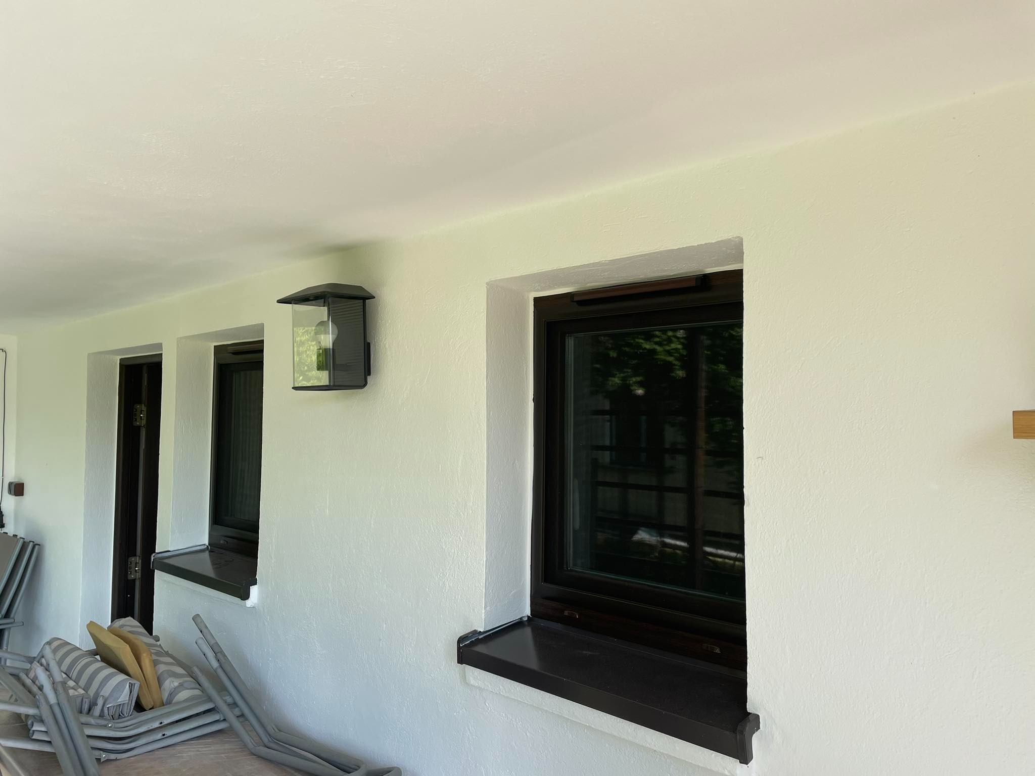 Świeżo pomalowana elewacja z oknami w ciemnych ramach i nowoczesną lampą zewnętrzną, widoczne złożone krzesła ogrodowe w tle.