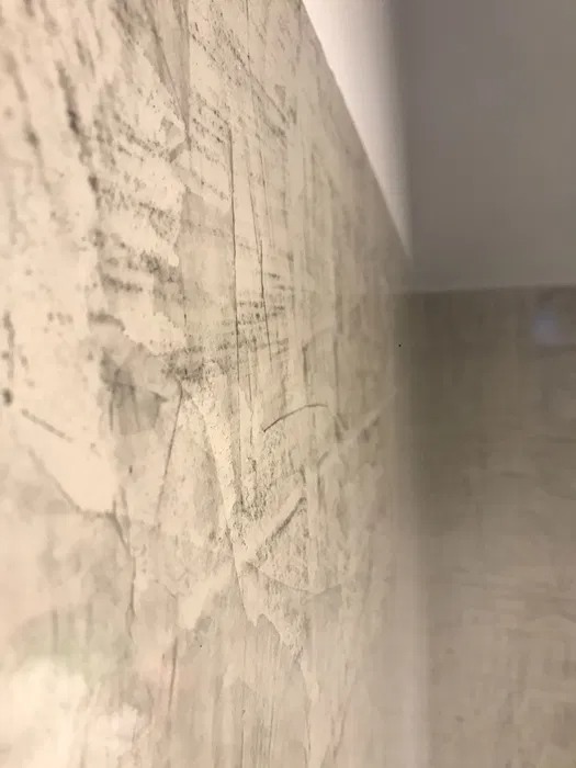 Szara ściana z fakturą betonu, widoczna z bliskiej perspektywy, zarysowania i nierówności na powierzchni, biały sufit w górnym rogu.