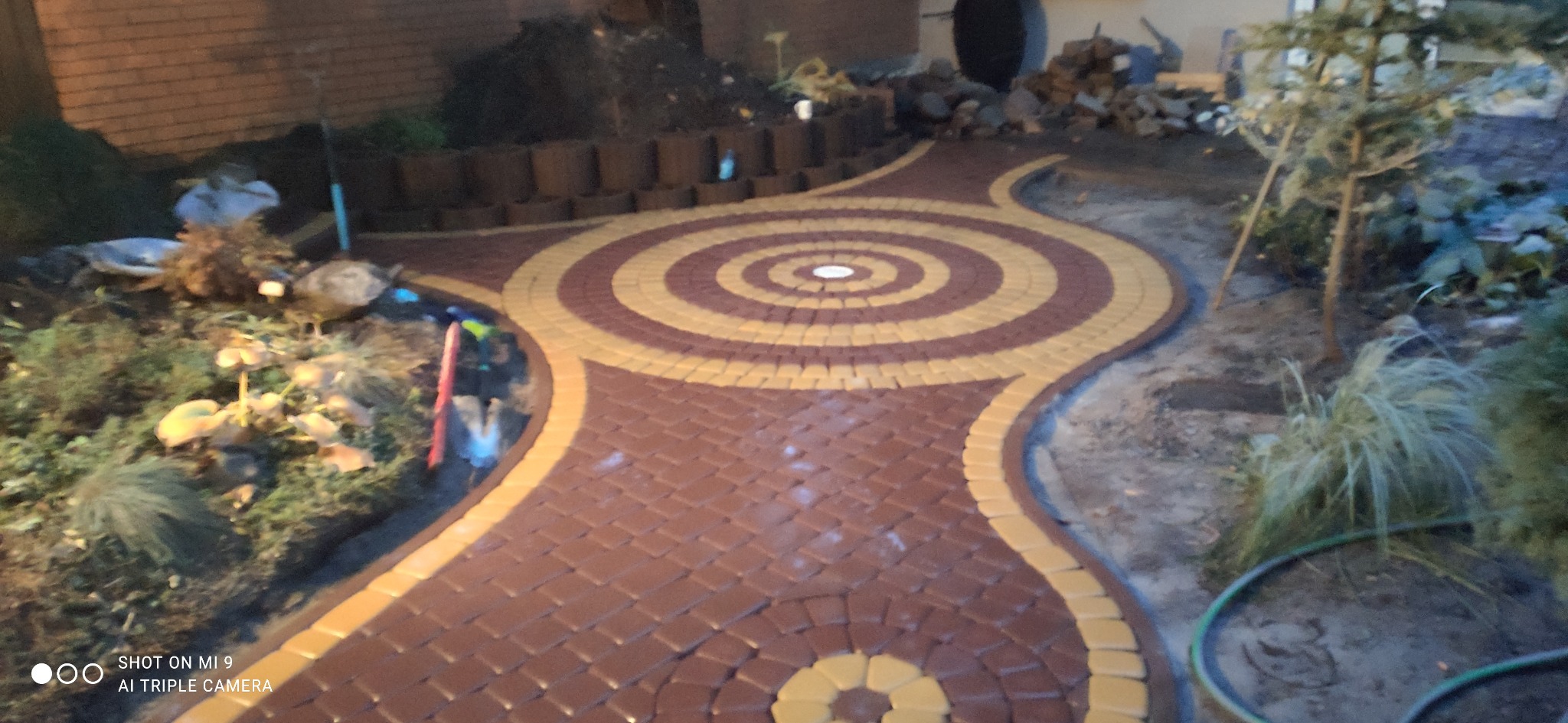 Okrągły wzór z brązowo-żółtej kostki brukowej tworzący ścieżkę w ogrodzie, widok z niskiej perspektywy, oświetlenie wieczorne.