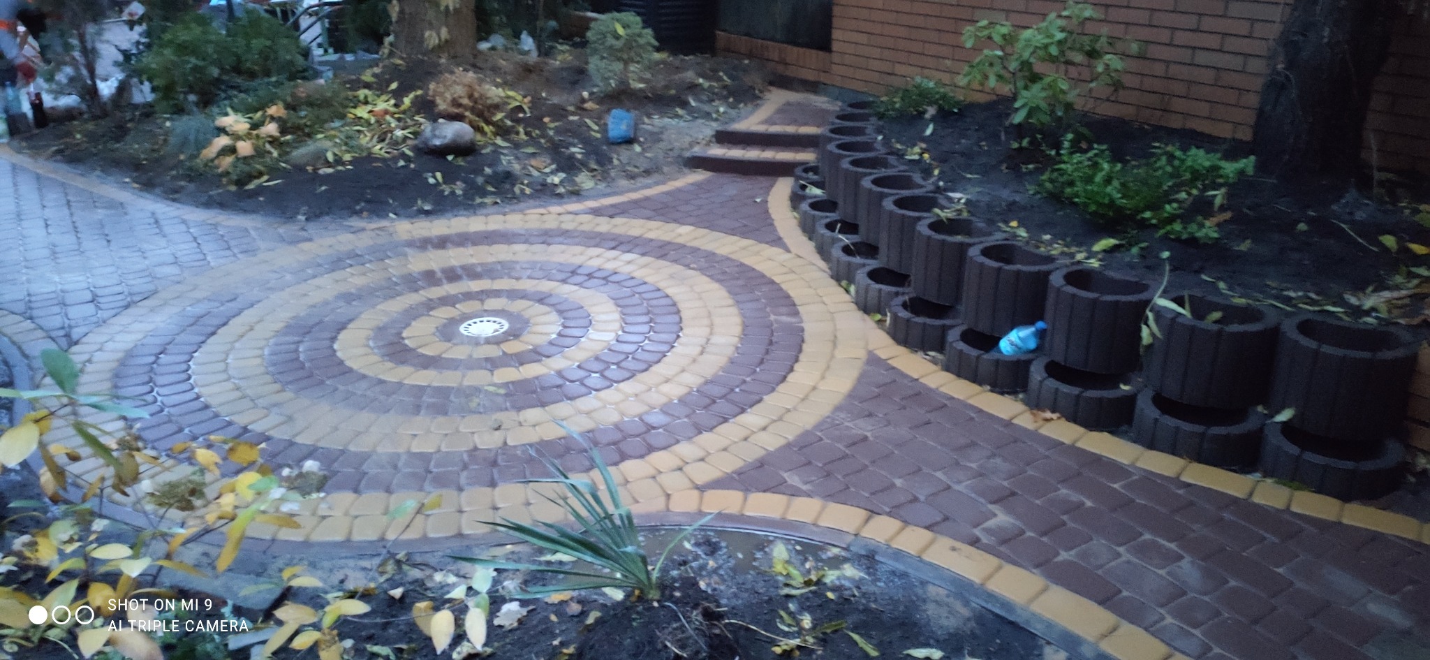 Ogród z okrągłym wzorem z kostki brukowej w odcieniach brązu i beżu, obrzeża z donic betonowych i schodki, widok z góry.