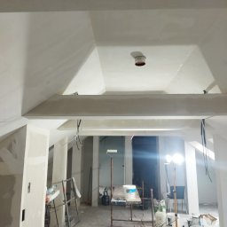 Bud - Poddasze w trakcie prac wykończeniowych: widoczne elementy konstrukcyjne, przygotowane ściany, narzędzia malarskie, oświetlenie robocze i wiadra z farbą.