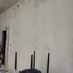 Bud - Ściana po nałożeniu pierwszej warstwy szpachli, widoczne niedoskonałości i otwory na gniazdka elektryczne, w tle framuga drzwi zabezpieczona niebieską taśmą malarską oraz fragment wiadra z zaprawą...