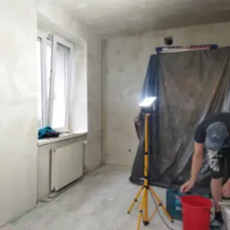 Pokój w trakcie szpachlowania ścian i sufitu, widoczna lampa na statywie, narzędzia i zabezpieczenie folią