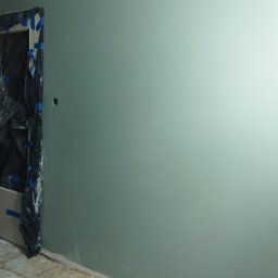 Bud - Świeżo pomalowana ściana w odcieniach szarości i zieleni z widocznymi otworami na gniazdka elektryczne, zabezpieczone folią ościeżnice drzwi i surowa podłoga ze zwiniętym kablem.