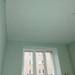 Pomalowane na jasnozielono ściany i sufit w pokoju z białym oknem, przez które widać budynek naprzeciwko.