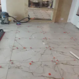 Podłoga z płytek imitujących marmur w trakcie układania, widoczne krzyżyki dystansowe, poziomica, odkurzacz przemysłowy i głośnik Bluetooth.