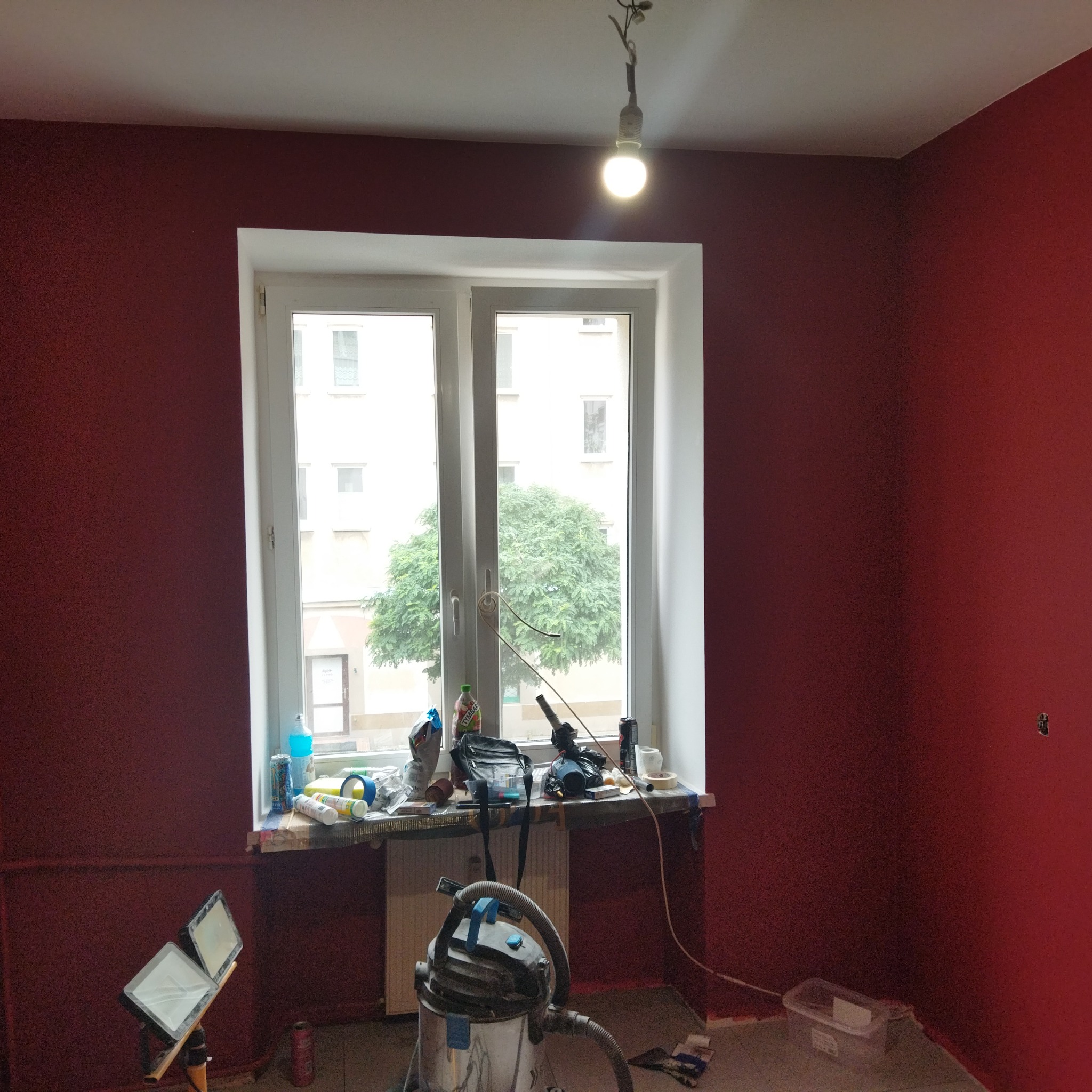 Pokój w trakcie malowania na burgundowo, z widocznymi narzędziami i sprzętem malarskim na parapecie i podłodze, oświetlony lampą roboczą.
