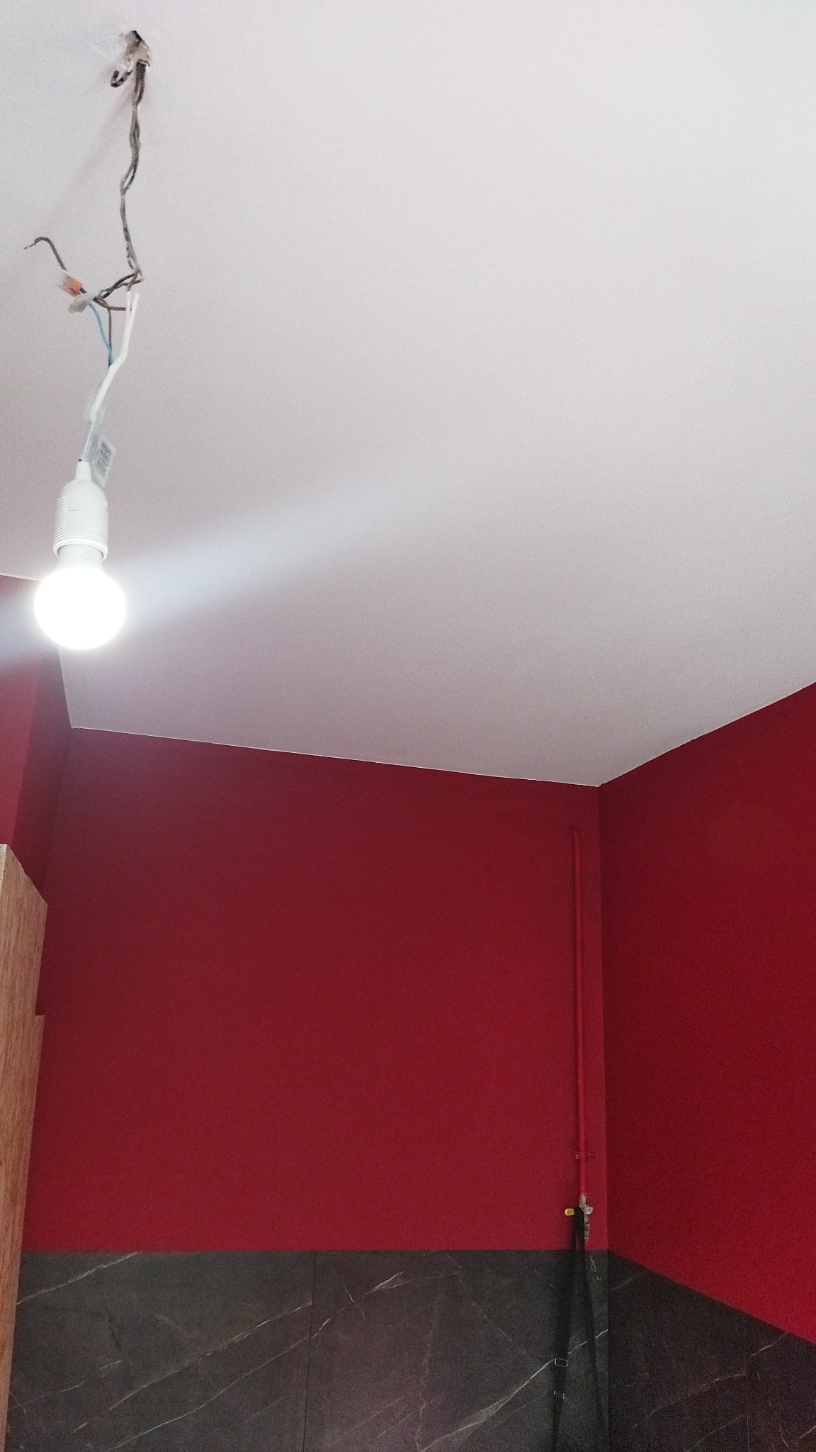 Czerwona ściana w pokoju z białym sufitem i odsłoniętymi kablami z żarówką, widoczna dolna część ściany wykończona płytkami imitującymi marmur.
