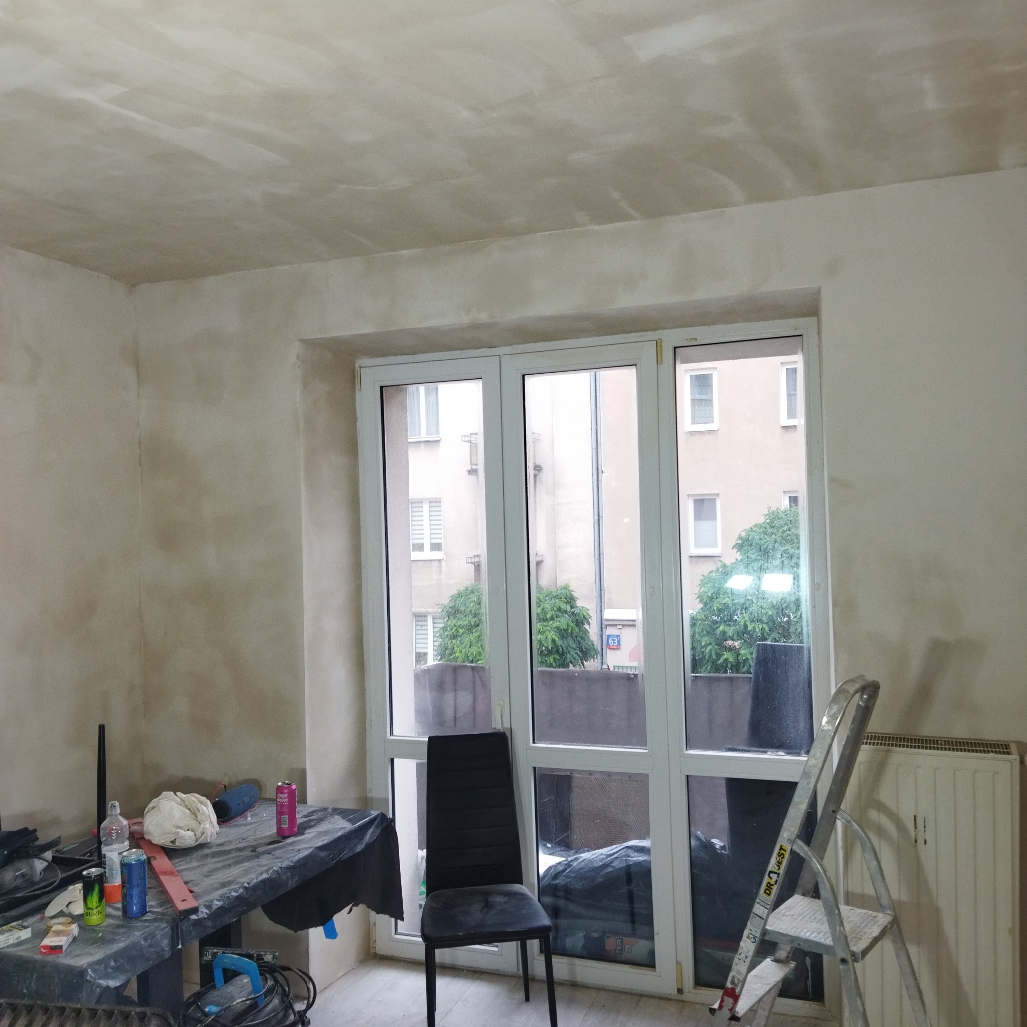 Pokój w trakcie prac wykończeniowych: ściany i sufit po nałożeniu warstwy podkładowej, widoczny stół z narzędziami i drabiną, okno z widokiem na budynek mieszkalny.