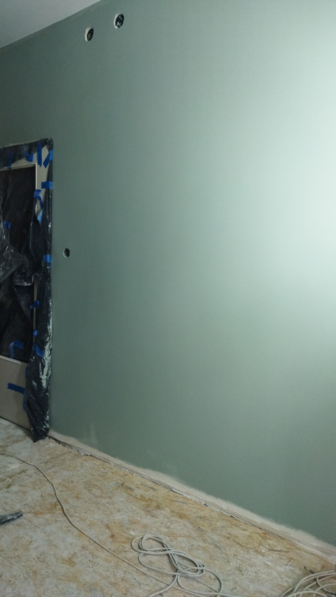 Świeżo pomalowana ściana w odcieniach szarości i zieleni z widocznymi otworami na gniazdka elektryczne, zabezpieczone folią ościeżnice drzwi i surowa podłoga ze zwiniętym kablem.