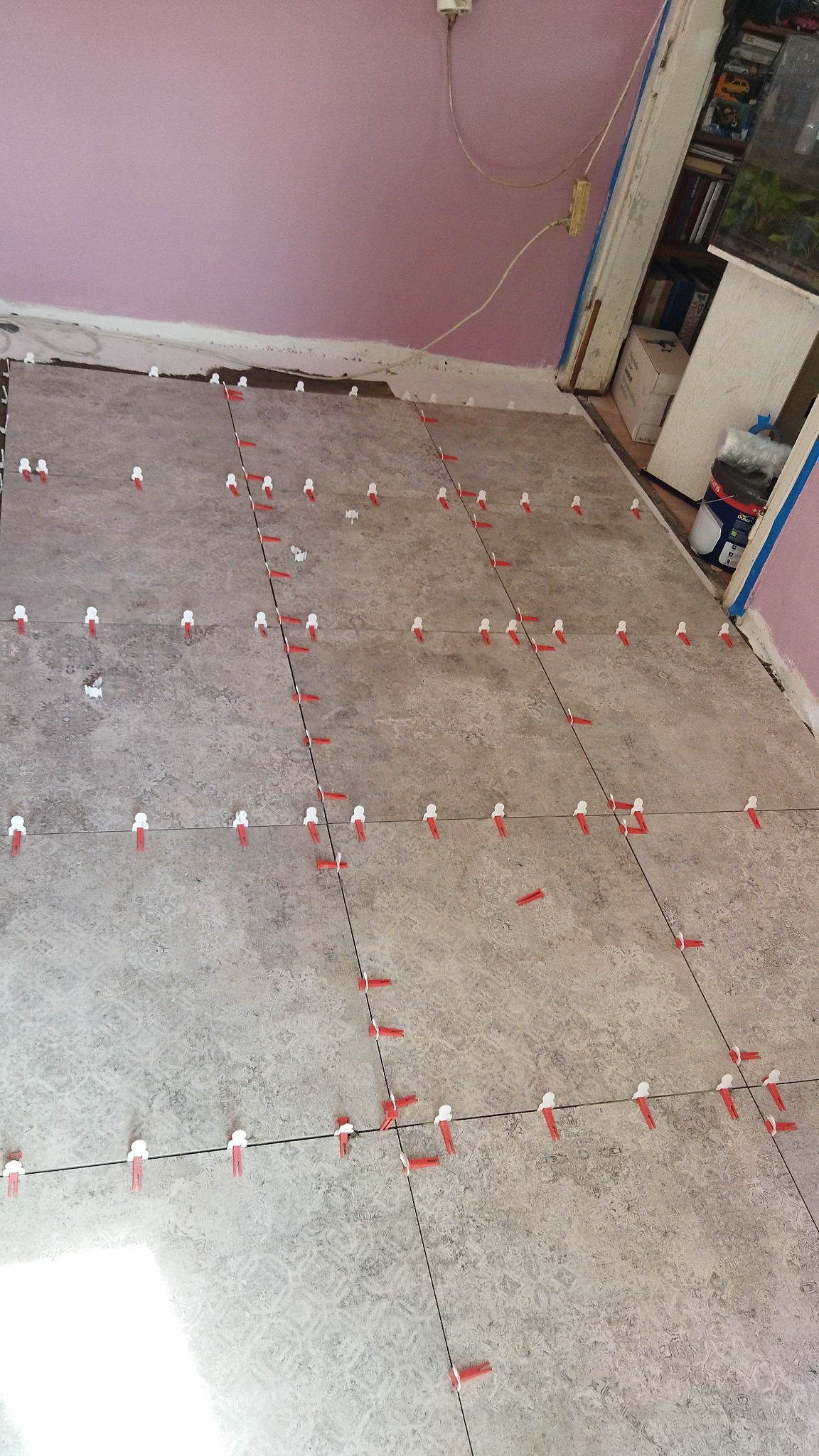 Szare płytki podłogowe w trakcie układania, z użyciem czerwonych i białych klipsów poziomujących, widoczne niedociągnięcia przy ścianie i różowa ściana w tle.