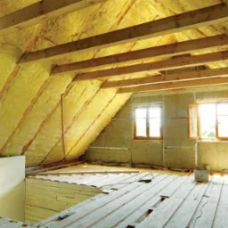 Poddasze w trakcie ocieplania wełną mineralną, widoczne drewniane belki konstrukcyjne, dwa okna z drewnianą stolarką i częściowo ułożona drewniana podłoga.