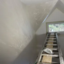 Ściana poddasza po nałożeniu dekoracyjnej, szarej gładzi, widoczna drabina oparta o okno z narzędziem do gładzi na szczeblu.