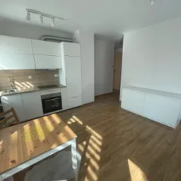 Jasne wnętrze mieszkania po remoncie: panele podłogowe, biała zabudowa kuchenna, stół z dekoracją w kształcie gwiazdy, komoda i oświetlenie punktowe.